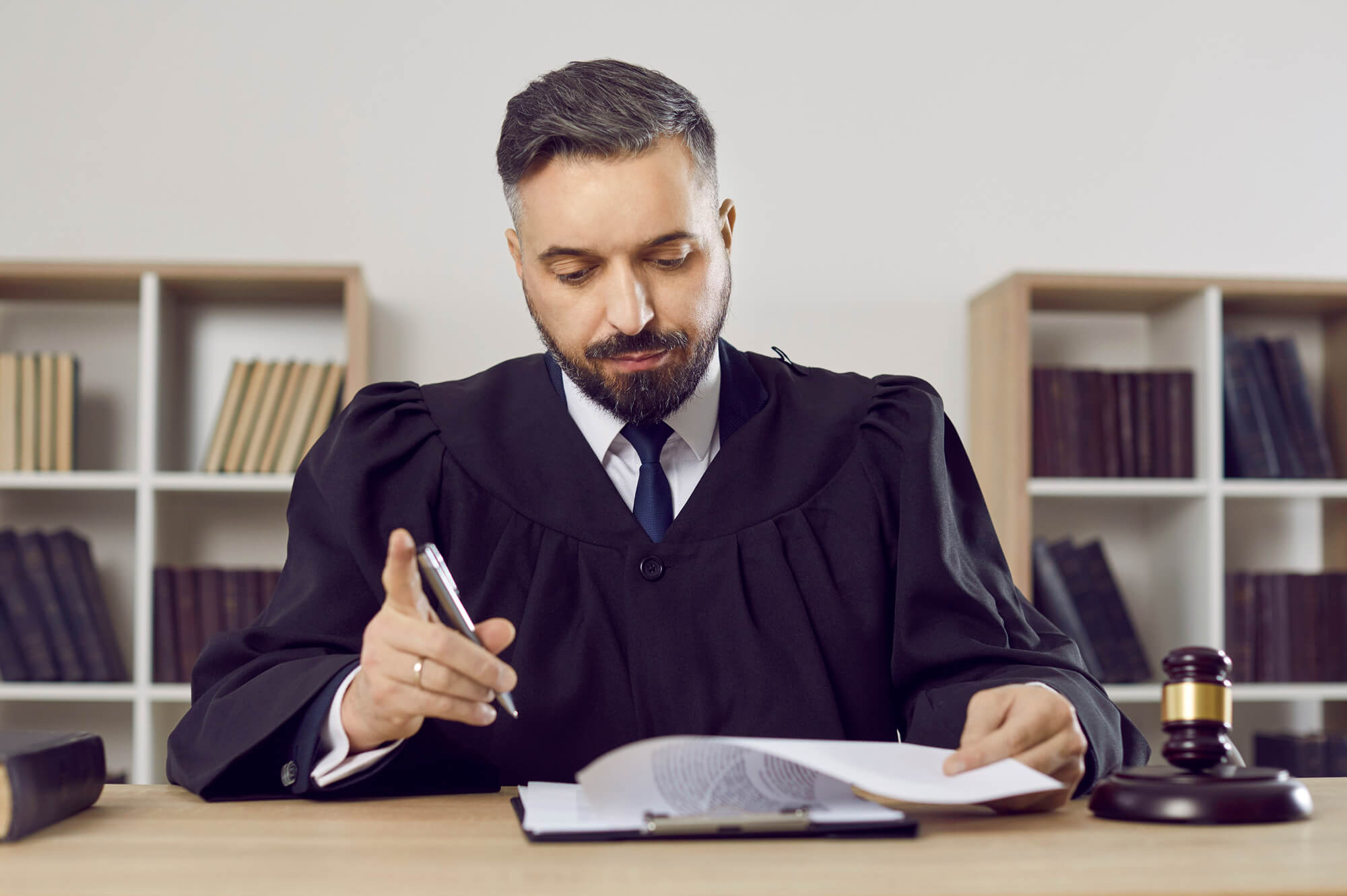 Ein Richter in einer Robe sitzt in seinem Arbeitszimmer am Tisch, darauf befindet sich ein Richterhammer. Der Richter sichtet Unterlagen für ein Gerichtsverfahren.