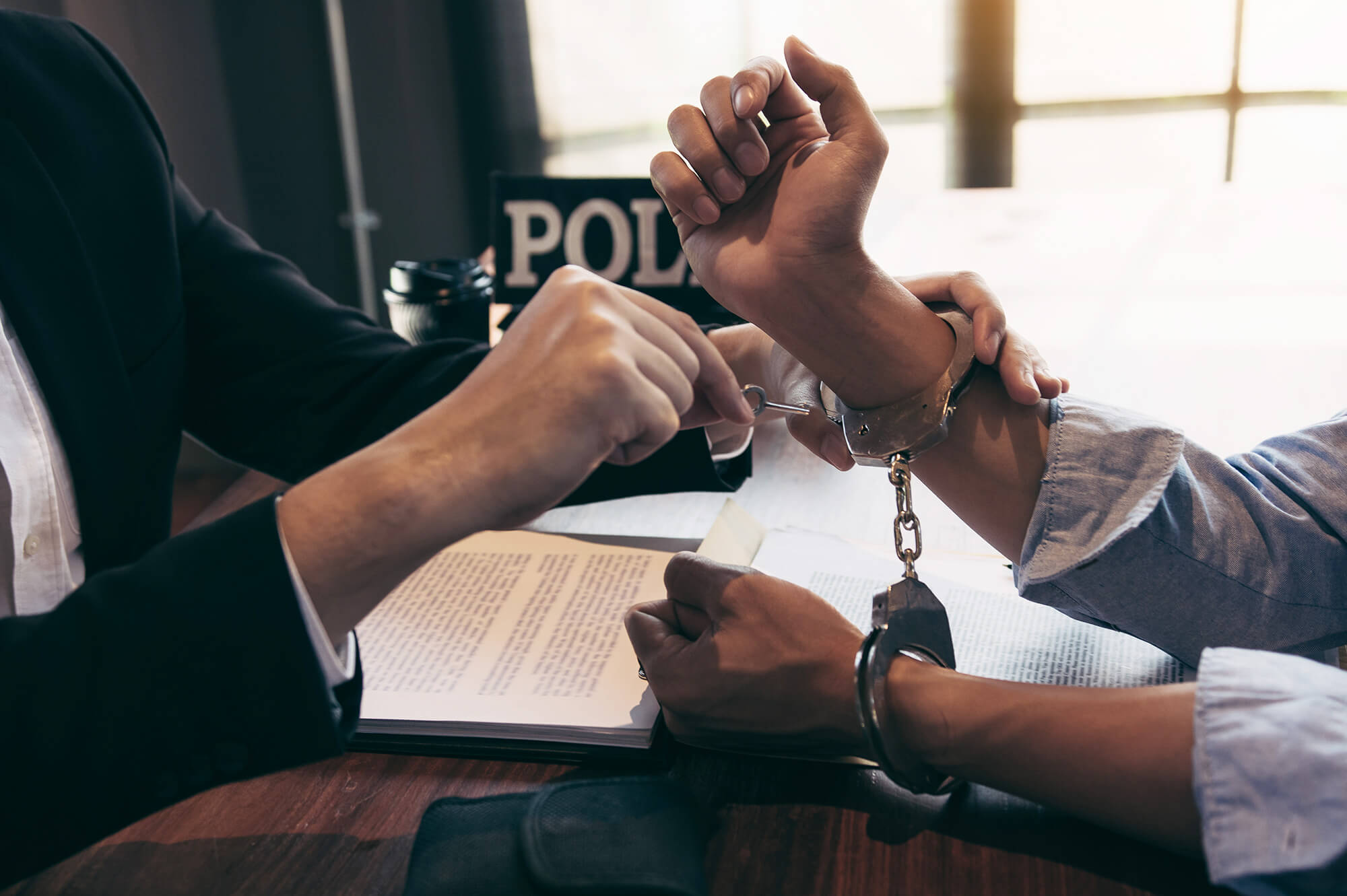 An einem Tisch sitzen ein Ermittler und ein Beschuldigter in Handschellen. Auf dem Tisch ist ein Schild mit Polizei. Der Ermittler schließt dem Beschuldigten die Handschellen auf.